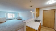 This single-floor unit is designed with a practical, open-concept layout that connects the kitchen and living areas into one cohesive space. The kitchen is outfitted with warm oak cabinetry providing ample storage, a white refrigerator, an electric range with microwave above, and a double-basin sink set into a light-colored countertop. A peninsula extends from the kitchen into the living area, creating a natural boundary while maintaining visual openness. A pendant light fixture accents the peninsula, adding a functional focal point to the shared space.

The living area is generously proportioned, featuring neutral carpet, white walls, and warm wood baseboards that run consistently throughout. Natural light enters through windows, and baseboard heating units are present along the perimeter walls. The layout, as visible in the floor plan model, includes dedicated bedroom spaces, each carpeted and featuring baseboard heat. One bedroom includes a wall-mounted air conditioning unit and a window with an exterior view.

Two full bathrooms are present, each featuring tile flooring in a neutral tone, a tub/shower combination, toilet, and a vanity with wood cabinetry. The overall aesthetic is clean and functional, with consistent use of light neutral tones, oak wood accents, and durable materials throughout.