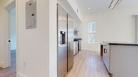 This contemporary single-floor residence offers a clean, modern aesthetic defined by crisp white walls, light wood-style flooring, and an abundance of natural light throughout. The open kitchen is a standout feature, equipped with full stainless steel appliances including a large side-by-side refrigerator with water dispenser, a stainless range with hood, and a dishwasher. Sleek cabinetry with dark countertops and a kitchen island provide ample prep and storage space, while a window above the range allows natural light to flow in. The living area is bathed in sunlight from large floor-to-ceiling sliding glass doors that open to views of the surrounding urban landscape, including tree-lined streets and neighboring buildings. The bedrooms are generously sized, each featuring light-filled windows and light wood-style flooring. One bedroom includes a walk-in closet with a built-in hanging rod. The unit features two bathrooms: one with a tub-shower combination and a backlit vanity mirror mounted above a floating white vanity with dark countertop, and a second with a stand-alone shower. Both bathrooms carry the same modern, minimalist design language with black hardware accents and matching flooring throughout.
