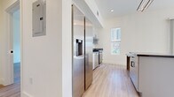 This contemporary single-floor residence offers a clean, modern aesthetic defined by crisp white walls, light wood-style flooring, and an abundance of natural light throughout. The open kitchen is a standout feature, equipped with full stainless steel appliances including a large side-by-side refrigerator with water dispenser, a stainless range with hood, and a dishwasher. Sleek cabinetry with dark countertops and a kitchen island provide ample prep and storage space, while a window above the range allows natural light to flow in. The living area is bathed in sunlight from large floor-to-ceiling sliding glass doors that open to views of the surrounding urban landscape, including tree-lined streets and neighboring buildings. The bedrooms are generously sized, each featuring light-filled windows and light wood-style flooring. One bedroom includes a walk-in closet with a built-in hanging rod. The unit features two bathrooms: one with a tub-shower combination and a backlit vanity mirror mounted above a floating white vanity with dark countertop, and a second with a stand-alone shower. Both bathrooms carry the same modern, minimalist design language with black hardware accents and matching flooring throughout.