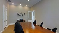 This single-floor office space offers a clean, professional environment well-suited for productivity and collaboration. The centerpiece of the space is a conference or meeting area anchored by a warm-toned hardwood conference table surrounded by ergonomic black office chairs, providing ample seating for meetings and discussions. The elevated ceilings, finished with crisp crown molding, give the space an open and airy feel, while recessed lighting ensures consistent, even illumination throughout.

The walls are finished in a neutral gray tone, contributing to a calm and focused atmosphere. A dedicated workstation area along one wall features a desk with a computer setup, ideal for individual work tasks. A decorative arrangement of circular wall art adds a stylish accent, bringing subtle character to the otherwise minimalist interior.

A white-framed glass-paned interior door connects to an adjacent room or hallway, allowing natural light to filter through while maintaining a sense of separation between areas. Light hardwood flooring runs throughout, complementing the warm tones of the conference table and tying the overall aesthetic together cohesively. The space balances functionality with thoughtful design, making it a practical and inviting professional environment.