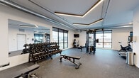 This modern fitness center offers a thoughtfully designed workout space that accommodates a wide range of exercise needs. The room is finished with durable speckled rubber flooring throughout, providing both traction and impact absorption ideal for high-intensity training. 

The ceiling features a striking geometric arrangement of linear LED lighting paired with exposed industrial-style ductwork, creating a contemporary aesthetic that balances functionality with style. Large floor-to-ceiling windows along multiple walls flood the space with natural light while offering expansive views of the surrounding area, enhancing the overall workout environment.

The strength training zone is well-appointed with a full rack of dumbbells spanning a wide weight range, multiple adjustable benches, and a multi-functional cable machine system suitable for a variety of resistance exercises. Additional specialty equipment, including a leg extension/curl machine and cardio options, rounds out the offering.

Full-length mirrors line one wall, maximizing the sense of space and allowing users to monitor their form during exercise. A wall-mounted television provides entertainment. Separate doors lead to adjacent rooms, suggesting access to changing facilities or restrooms. The overall ambiance is clean, bright, and professional, making this a well-rounded fitness facility.