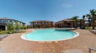This expansive outdoor amenity space is designed around a generously sized, freeform swimming pool with clear blue water and a surrounding stamped-concrete pool deck. Lounge chairs line the perimeter, offering ample space for sunbathing beneath open skies. An attached spa or wading area is integrated near the pool's edge, enhancing the resort-like atmosphere.

Adjacent to the pool, a substantial covered patio is constructed with natural stone masonry columns and matching exterior walls, lending the space a refined yet relaxed character. This shaded lounge area features mounted flat-screen televisions and comfortable wicker sectional seating with bold blue cushions, creating a functional outdoor living environment. The stamped paver flooring extends throughout, connecting multiple zones seamlessly.

Additional outdoor seating areas include modern metal dining tables with bench seating, positioned along the covered perimeter with direct views toward the pool and surrounding landscaping. Cornhole boards and open patio space provide casual recreation opportunities. Large black-framed floor-to-ceiling glass doors and windows connect the outdoor patio to an interior clubhouse space, ensuring a fluid indoor-outdoor flow. Mature palm trees, manicured planting beds, and the multi-story residential buildings in the background complete the well-appointed setting.