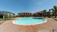 This expansive outdoor amenity space is designed around a generously sized, freeform swimming pool with clear blue water and a surrounding stamped-concrete pool deck. Lounge chairs line the perimeter, offering ample space for sunbathing beneath open skies. An attached spa or wading area is integrated near the pool's edge, enhancing the resort-like atmosphere.

Adjacent to the pool, a substantial covered patio is constructed with natural stone masonry columns and matching exterior walls, lending the space a refined yet relaxed character. This shaded lounge area features mounted flat-screen televisions and comfortable wicker sectional seating with bold blue cushions, creating a functional outdoor living environment. The stamped paver flooring extends throughout, connecting multiple zones seamlessly.

Additional outdoor seating areas include modern metal dining tables with bench seating, positioned along the covered perimeter with direct views toward the pool and surrounding landscaping. Cornhole boards and open patio space provide casual recreation opportunities. Large black-framed floor-to-ceiling glass doors and windows connect the outdoor patio to an interior clubhouse space, ensuring a fluid indoor-outdoor flow. Mature palm trees, manicured planting beds, and the multi-story residential buildings in the background complete the well-appointed setting.