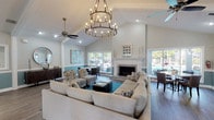 This well-appointed clubroom offers an inviting, single-floor layout designed for both relaxation and social gatherings. The main lounge area commands attention with soaring vaulted ceilings, a striking two-tiered candelabra chandelier, and rich dark hardwood-style flooring that extends throughout the space. A classic white-mantled fireplace serves as a central focal point, flanked by large sliding glass doors and windows that flood the room with natural light and provide views of the outdoor pool area. The lounge is furnished with a generous sectional sofa, patterned accent chairs, and a teal area rug that adds a pop of coastal color, complementing the soft blue wainscoting that runs along the lower walls. Adjacent to the main lounge, a dedicated dining area accommodates groups comfortably, while a built-in bar counter with dark bar stools provides a casual seating option. The fully equipped kitchen features white cabinetry, granite countertops, and stainless steel appliances, including a refrigerator, dishwasher, and microwave. A separate workspace or business center area is also visible, offering functional amenities for residents. Recessed lighting and ceiling fans throughout ensure a comfortable, well-lit environment year-round. The overall ambiance is bright, modern, and welcoming.