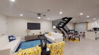 This two-floor space is designed with a lively, eclectic aesthetic that balances comfort and functionality. The main living area is a standout, featuring recessed lighting, a ceiling fan, and a wall-mounted flat-screen television. Bold yellow floral armchairs contrast a crisp white sofa, anchored by a vivid blue area rug that adds a striking pop of color. A sleek black staircase with vertical railing detail connects the floors, serving as an architectural focal point.

The kitchen is well-appointed with white shaker cabinetry, dark mosaic backsplash, granite-topped counters, and a full suite of stainless steel appliances. A breakfast bar with corrugated metal siding and red barstools adds industrial charm. Colorful floral curtains frame the space with personality.

The primary bedroom carries a coastal theme, with a woven rattan headboard, nautical bedding, and beach-inspired wall art, complemented by wood-look flooring and a walk-in closet with an adjacent vanity area.

The kids' bedroom is fun and functional, housing a twin bed alongside a house-shaped bunk bed, offering ample sleeping capacity. The bathroom features subway tile with a decorative mosaic accent band. Ceiling fans are present throughout, and the overall palette is bright and welcoming.