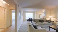 This single-floor residence showcases an open-concept layout that seamlessly connects the living room, dining area, and kitchen into one cohesive and functional space. The living area is carpeted and comfortably sized, accommodating a neutral-toned sectional sofa arrangement accented with decorative pillows. Framed artwork adorns the light-colored walls, adding a personal touch to the space. Adjacent to the living room, the dining area features a dark-toned table and chair set positioned near a large window that fills the space with natural light. The kitchen is situated just beyond the dining area and is equipped with white cabinetry, a refrigerator, and a countertop that appears to serve as a casual bar or prep area. Overhead flush-mount lighting provides consistent illumination throughout the open space. The neutral color palette of whites and beiges across the walls, carpet, and cabinetry creates a clean, cohesive aesthetic. Multiple windows throughout the space ensure ample natural light, contributing to an airy and welcoming atmosphere throughout this well-designed single-level home.