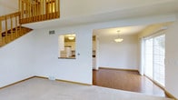 This thoughtfully designed multi-level residence spans three floors, offering a versatile and functional layout suited for comfortable everyday living. The main living area features plush carpeting and flows seamlessly into an adjoining space with warm hardwood flooring, creating a natural distinction between zones while maintaining an open, airy feel. A pass-through window connects the living area to the kitchen, which is outfitted with white cabinetry, a stone-style countertop, and standard appliances including a refrigerator. Pendant lighting fixtures illuminate both the kitchen and the adjoining dining or secondary living area, adding a welcoming ambiance throughout. Crisp white walls and clean lines give the interior a bright, neutral palette that is easy to personalize. A loft-style upper level is visible from the main floor, accented by a classic wood railing that adds architectural character and visual interest to the vertical space. HVAC vents are thoughtfully integrated into the walls and floors, ensuring year-round comfort. The three-floor layout provides ample room for living, working, and entertaining, with each level offering its own distinct functionality within a cohesive overall design.