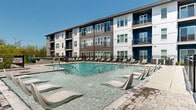 Located at 5700 Crossings Blvd in Nashville, TN, this outdoor amenity area is designed to deliver a resort-style experience for residents. The centerpiece is a generously sized swimming pool fitted with built-in in-water chaise loungers, allowing residents to relax directly in the water. The surrounding pool deck is finished with clean, light-toned concrete and pavers, and is lined with rows of modern sling-style lounge chairs, providing ample space for sunbathing under the open Tennessee sky. The pool area is enclosed by sleek black iron fencing, offering both security and an unobstructed view of the surrounding landscape and multi-story residential buildings, which feature a contemporary exterior palette of white, dark wood-tone siding, and bold navy blue accents. Adjacent to the pool is a covered outdoor patio lounge area, distinguished by a warm wood-paneled ceiling, ceiling fan, and exposed brick columns. This space is furnished with round dining tables, chairs, and plush seating arrangements, creating an inviting setting for both casual gatherings and quiet relaxation. Together, these amenities establish a cohesive, upscale outdoor living environment suited to the Nashville climate.
