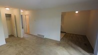This single-floor space presents a straightforward and functional layout designed to maximize usability. The main living area is finished with wall-to-wall carpeting in a neutral beige tone, contributing to a warm and understated atmosphere throughout. The walls are painted in a clean, off-white finish, creating a bright and open feel despite the absence of large windows visible in the primary living zone. A floor-level HVAC vent is positioned along the base of the central dividing wall, indicating a built-in climate control system integrated into the unit's infrastructure. The layout features a central partial wall that separates the main living space from an adjoining room, which also features matching carpet and a ceiling light fixture, suggesting use as a dining area or secondary living space. A hallway extending from the left side of the main room leads to additional areas, including what appears to be a bathroom or utility space, as indicated by the door and interior lighting visible through the opening. Ceiling-mounted light fixtures provide ambient illumination throughout. Overall, the space offers a clean, neutral canvas with a connected and practical floor plan.