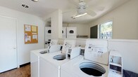 This single-story shared laundry facility is designed for convenient, communal use. The room features a row of top-loading washing machines positioned along the front, paired with multiple stacked dryer units arranged along the back wall, providing ample capacity for residents. The white appliances and bright white walls give the space a clean, utilitarian appearance.

A ceiling fan is centrally mounted overhead, helping to circulate air and maintain comfortable conditions during use. Natural light enters through a window on the far wall, offering a view of greenery outside and contributing to the room's open, airy feel. A structural column adds a subtle architectural element to the interior layout.

Practical additions include a cork bulletin board mounted on the wall near the entrance, which serves as a central point for posting notices or instructions. A chalkboard on the adjacent wall provides another communication surface. The facility is straightforward and functional, with a layout that allows users to move easily between machines, making it an efficient and well-organized shared laundry space.