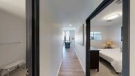 This single-floor residence showcases a thoughtfully designed modern interior with a cohesive aesthetic defined by clean lines, white walls, and light hardwood-style flooring that flows throughout the main areas. Bold black door frames and trim provide striking contrast against the bright, neutral palette, lending the space a contemporary edge.

The kitchen is well-appointed with dark granite countertops, two-tone cabinetry featuring white lower units and textured gray upper cabinets, a white subway tile backsplash, and stainless steel appliances including a range and full-size refrigerator. A pendant light fixture adds a decorative touch above the counter space.

The hallway connects the kitchen and living areas to the private bedroom, with framed artwork displayed along the wall. The living area benefits from large windows fitted with roller shades, offering urban views and ample natural light. The open, comfortable layout accommodates a full seating arrangement centered around a wall-mounted television.

The bathroom is designed with accessibility in mind, featuring a wall-mounted sink, an illuminated backlit mirror, light tile flooring, grab bars, and an open roll-in shower area with a bench — combining practicality with a clean, minimalist aesthetic.