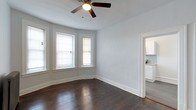 This single-floor space showcases a thoughtfully designed layout with classic architectural details and functional living areas. The main living area is highlighted by rich, dark hardwood flooring that extends throughout, providing a warm contrast to the light gray walls. A curved bay window configuration allows natural light to fill the room, featuring two windows with white trim that complement the clean, neutral color palette. A ceiling fan with an integrated light fixture is centrally mounted, offering both air circulation and ambient lighting.

White painted baseboards and door trim add a crisp, traditional finish to the space, while the smooth ceiling maintains a clean, uncluttered aesthetic. An open doorway leads directly into the kitchen, which is visible from the main living area and features white cabinetry, a tiled backsplash with a subtle pattern, and what appears to be stone or granite countertops. The kitchen continues the hardwood flooring, creating a cohesive flow between spaces. Overall, the space presents a balance of classic architectural character and practical design, well-suited for comfortable everyday living.