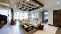This sophisticated clubhouse is a masterfully designed communal space that blends modern architecture with refined interior details. The open-plan layout creates a seamless flow throughout the space, anchored by a generously sized lounge area furnished with plush, tufted cream sectional sofas, dark espresso accent tables, and carefully curated decorative accessories that lend a polished, upscale feel.

One of the most striking architectural features is the ceiling design, which showcases bold dark wood beam detailing set against a crisp white backdrop, complemented by recessed lighting that evenly illuminates the space. Large floor-to-ceiling windows line the perimeter, flooding the interior with natural light and offering clear views of the surrounding outdoor areas and landscaping.

Light-toned large-format tile flooring extends throughout, enhancing the sense of openness and continuity. A textured stone column serves as a natural divider, adding visual depth while defining separate functional zones within the space. In the background, what appears to be a bar or beverage service area is visible, pointing to the clubhouse's role as a complete social hub.

The overall ambiance is one of contemporary elegance — inviting yet refined, and well-suited for both casual relaxation and social gatherings.