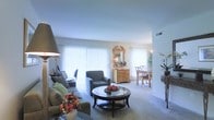 This single-floor residence features an open-plan layout that seamlessly connects the living and dining areas, creating a sense of spaciousness and easy flow throughout. The living area is carpeted and comfortably appointed, centered around a round glass-topped coffee table with a dark wood base. Plush upholstered armchairs and a sofa provide ample seating, while a large floor lamp adds warmth to the space. Floor-to-ceiling vertical blinds along the expansive windows allow generous amounts of natural light to fill the room, giving the interior a bright, airy quality.

Transitioning naturally from the living area, the dining space features a wooden table and chairs alongside an ornate wooden dresser topped with a decorative mirror, lending a classic, traditional character to the space. A potted plant and floral arrangements throughout add organic touches of color and life. The neutral tones of the walls and carpet create a cohesive, understated backdrop that ties the open-plan space together. Overall, the layout and design prioritize comfort, light, and a welcoming, livable ambiance.