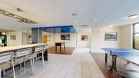 This single-floor amenity space is designed as a dynamic, multi-use lounge that blends recreation and productivity. The open layout is defined by a combination of light tile flooring and rich hardwood sections, clearly delineating different activity zones. A distinctive blue painted ceiling panel serves as an architectural focal point, adding a bold visual element to the otherwise neutral white walls and ceiling with recessed lighting throughout.

The recreational zone features a foosball table and a full ping-pong table, both positioned on the hardwood flooring area, while multiple wall-mounted televisions provide additional entertainment options. Framed artwork and a large, vibrant photographic mural depicting a theater marquee contribute to the space's energetic, urban character.

A co-working area includes a long communal high-top table with bar-height seating, accented by pendant lighting overhead. Adjacent to this, a separate lounge zone features bright chartreuse accent walls, wall-mounted screens, and a built-in counter with additional seating, flooded with natural light from large windows overlooking an outdoor courtyard. The overall ambiance is modern and lively, designed to encourage both social interaction and focused work.