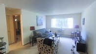 This single-floor space features a living room designed for comfort and functionality. The room is finished with wall-to-wall carpeting in a neutral tone, contributing to a warm and inviting atmosphere. Light-colored walls keep the space feeling open and airy, complemented by a large window that allows generous natural light to fill the room while offering a view of the surrounding greenery outside.

The living area is furnished with a blue sofa accented by decorative pillows, paired with a patterned accent chair that adds visual interest to the space. A round glass-top coffee table serves as a central focal point, flanked by side tables with a decorative lamp. A wall-mounted piece of artwork adds a subtle touch of color to the otherwise neutral palette.

Climate control is provided by a wall-mounted AC unit positioned beneath the window, with a baseboard heating unit running along the exterior wall — offering year-round comfort. The layout is straightforward and efficient, making practical use of the available space. Overall, the living room presents a cozy, well-lit environment suitable for everyday living.
