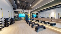 Located at 3615 Tryclan Drive in Charlotte, NC, this fitness studio is a thoughtfully designed group exercise space that combines functionality with a vibrant, energizing aesthetic. The single-floor layout offers a well-organized open floor plan finished with light wood-tone flooring that brightens the room and provides a clean, modern feel underfoot.

The studio features exposed ductwork and industrial-style ceilings with ceiling fans and track lighting, lending an urban edge to the space. A full-length illuminated mirror wall runs along one side, amplifying the sense of space and allowing exercisers to monitor their form. Warm LED accent lighting beneath the mirror adds an inviting, polished touch.

The room is equipped with multiple step platforms arranged in rows for group classes, along with dual dumbbell racks offering a range of free weights and TRX suspension straps mounted to the wall. A colorful rack of stability balls adds both visual interest and versatility to the available equipment. Large-format nature-inspired wall murals serve as bold focal points, creating an immersive and motivating atmosphere.

Floor-to-ceiling windows along one wall flood the space with natural light and provide views of the surrounding outdoor courtyard. A glass-paneled door connects the studio to adjacent fitness areas, enhancing flow and accessibility. A ceiling-mounted flat-screen display further supports guided workout sessions, making this studio well-suited for a variety of fitness classes.
