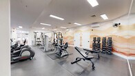Located at 8732 Town and Country Blvd in Ellicott City, MD, this fitness center offers a comprehensive workout environment across a generously sized single-floor layout. The space is thoughtfully organized into distinct workout zones designed to accommodate a wide range of fitness activities.

The cardio area features a full row of Spirit treadmills and upright stationary bikes positioned alongside large windows that allow natural light to fill the room and provide views of the outdoors. A floor-to-ceiling mirror along one wall amplifies the sense of openness throughout this section.

The strength training area is equipped with multiple cable and selectorized machines, a power rack loaded with a barbell, adjustable benches, and a well-stocked dumbbell rack — providing options for both beginners and experienced lifters. Rubber flooring mats are placed strategically beneath the equipment to ensure safety and durability.

A standout design element is the warm, desert-inspired wave mural adorning the accent wall in the weight room, adding an artistic and inviting atmosphere to the space. Open shelving units offer additional storage throughout the facility.

Bright recessed panel lighting throughout the ceiling ensures consistent, even illumination across all areas. The clean white walls, neutral flooring, and organized equipment layout contribute to a professional and welcoming fitness environment.