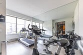 Located at 446 Kerr St in Oakville, Ontario, this dedicated fitness room offers a well-appointed workout space designed for functionality and comfort. The room features light hardwood-style flooring and crisp white walls that contribute to a clean, modern aesthetic throughout.

Natural light pours in through a bank of large windows fitted with roller shades, offering views of the surrounding Oakville streetscape. Floor-to-ceiling mirrors line one wall, visually expanding the space and providing users with proper form feedback during exercise.

The room is equipped with a diverse range of fitness equipment, including a treadmill, rowing machines, a stationary bike, a free weights rack with dumbbells, and an adjustable workout bench. A wall-mounted flat-screen television is positioned for easy viewing from multiple workout stations, adding an entertainment element to training sessions.

Entry is provided through a striking black-framed glass door, which connects the fitness room to an adjacent common area, allowing natural light to flow through while maintaining a sense of separation. Informational postings and a hand sanitizer station near the entrance reflect a thoughtful, user-focused design. A stainless steel waste bin completes the tidy, well-maintained environment. This fitness room delivers a comprehensive workout facility in a compact, efficiently organized layout.