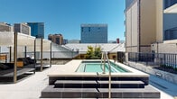 This outdoor amenity space is a well-appointed urban retreat situated within the courtyard and deck areas of a contemporary multi-story residential building. The centerpiece of the space is a clean, modern swimming pool with a tiled surround, complemented by a raised spa that offers a relaxing escape above the pool deck level. The pool area is finished with large-format concrete pavers and bordered by sleek black metal railings, providing safety while maintaining open sightlines throughout.

Surrounding the pool, the deck extends into dedicated lounging zones equipped with canopied daybeds and seating, ideal for outdoor relaxation. The space benefits from abundant natural light throughout the day thanks to its open-sky design. Safety features, including a life ring station and emergency shut-off switch, are thoughtfully integrated into the layout.

The surrounding multi-story building façade, clad in warm brick and neutral tones with private balconies overlooking the amenity area, creates an enclosed yet airy courtyard atmosphere. String lights are strung between structures, adding an inviting ambiance during evening hours. Panoramic views of the downtown skyline and distant mountain range are visible from the deck, offering a striking urban backdrop that elevates the overall experience of the space.