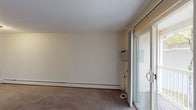 This single-floor unit at 1585 Smith St in North Providence, Rhode Island, offers a straightforward, functional layout across several well-defined spaces. The living area is generously sized, featuring neutral-toned carpet flooring, cream-painted walls, and baseboard heating that runs along the perimeter. A sliding glass door opens directly onto a private balcony, bringing in natural light and views of surrounding greenery. A wall-mounted air conditioning unit provides additional climate control.

A second carpeted room serves as a bedroom, equipped with its own baseboard heating, a ceiling-mounted light fixture, and a window framing outdoor tree views. A closet with a standard rod provides built-in storage. The room maintains the same clean, neutral aesthetic found throughout the unit.

The kitchen features warm wood cabinetry, dark granite-style countertops, wood-look vinyl flooring, and stainless steel appliances including a full-size refrigerator. The layout is compact and efficient, offering practical workspace.

The bathroom is cleanly finished with light ceramic tile flooring, a white vanity with ample cabinet storage, a mirrored medicine cabinet, a multi-bulb vanity light fixture, and a full bathtub with light blue tile surround. The overall ambiance of the unit is clean, neutral, and move-in ready.