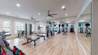 The HealthHUB Fitness Center is a spacious, single-floor facility designed to support a comprehensive workout experience. The interior features warm hardwood-style flooring throughout, crisp white walls, and recessed lighting complemented by multiple ceiling fans that maintain airflow and comfort during exercise sessions.

The main workout area is generously sized and thoughtfully organized, housing a diverse selection of strength training equipment including a multi-station cable machine, adjustable weight benches, a power tower, and a stair climber. A full-length wall mirror expands the sense of space and allows members to monitor their form. Cardio equipment such as treadmills, ellipticals, and a stationary cycle are arranged to maximize floor space and ease of movement.

A dedicated functional training zone features a HealthHUB-branded stair sled and a stretch mat area, while a rack of kettlebells and additional free weights lines the wall. Natural light flows in through multiple windows, offering views of the surrounding outdoor area, including what appears to be a pool. A motivational wall decal reinforces the fitness-focused atmosphere.

A utility corridor provides added convenience with a water fountain, built-in shelving, a granite-topped vanity counter, and sanitation stations, ensuring members have everything they need for a clean, comfortable visit.