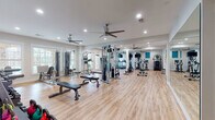 The HealthHUB Fitness Center is a spacious, single-floor facility designed to support a comprehensive workout experience. The interior features warm hardwood-style flooring throughout, crisp white walls, and recessed lighting complemented by multiple ceiling fans that maintain airflow and comfort during exercise sessions.

The main workout area is generously sized and thoughtfully organized, housing a diverse selection of strength training equipment including a multi-station cable machine, adjustable weight benches, a power tower, and a stair climber. A full-length wall mirror expands the sense of space and allows members to monitor their form. Cardio equipment such as treadmills, ellipticals, and a stationary cycle are arranged to maximize floor space and ease of movement.

A dedicated functional training zone features a HealthHUB-branded stair sled and a stretch mat area, while a rack of kettlebells and additional free weights lines the wall. Natural light flows in through multiple windows, offering views of the surrounding outdoor area, including what appears to be a pool. A motivational wall decal reinforces the fitness-focused atmosphere.

A utility corridor provides added convenience with a water fountain, built-in shelving, a granite-topped vanity counter, and sanitation stations, ensuring members have everything they need for a clean, comfortable visit.