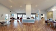 This single-floor space showcases a striking open-concept design centered around a dramatic floor-to-ceiling double-sided fireplace clad in a textured white panel surround. The fireplace serves as the architectural focal point of the layout, rising from a clean, low-profile base to a towering column that commands attention from every angle of the room.

The expansive main area features warm-toned hardwood flooring that extends throughout, complementing the bright white ceiling outfitted with recessed lighting. Soaring ceiling heights amplify the sense of openness and volume across the space.

A lounge area is positioned to one side, furnished with modern accent chairs, a low-profile sofa, and a bold red table, creating a stylish conversational setting. Large windows and glass doors line the perimeter walls, flooding the interior with natural light and offering views to the outdoors.

On the opposite side, a bar or kitchenette area is visible, equipped with counter seating, pendant lighting, and cabinetry, providing a functional complement to the social atmosphere. The overall ambiance is sleek, contemporary, and welcoming, making this space well-suited for entertaining or communal use.