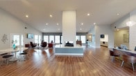 This single-floor space showcases a striking open-concept design centered around a dramatic floor-to-ceiling double-sided fireplace clad in a textured white panel surround. The fireplace serves as the architectural focal point of the layout, rising from a clean, low-profile base to a towering column that commands attention from every angle of the room.

The expansive main area features warm-toned hardwood flooring that extends throughout, complementing the bright white ceiling outfitted with recessed lighting. Soaring ceiling heights amplify the sense of openness and volume across the space.

A lounge area is positioned to one side, furnished with modern accent chairs, a low-profile sofa, and a bold red table, creating a stylish conversational setting. Large windows and glass doors line the perimeter walls, flooding the interior with natural light and offering views to the outdoors.

On the opposite side, a bar or kitchenette area is visible, equipped with counter seating, pendant lighting, and cabinetry, providing a functional complement to the social atmosphere. The overall ambiance is sleek, contemporary, and welcoming, making this space well-suited for entertaining or communal use.