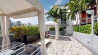 This single-level outdoor space presents a well-appointed courtyard retreat with a distinctly tropical, resort-inspired design. The centerpiece of the space is a series of white fabric cabanas with flowing curtains, providing shaded lounging areas ideal for relaxation and social gatherings. Beneath these canopies, contemporary gray outdoor seating with accent cushions offers a comfortable and stylish setting for guests.

The courtyard is finished with a patterned paver surface that adds visual texture underfoot while complementing the clean, modern aesthetic of the surrounding architecture. Tall, manicured hedges in sleek black planters serve as privacy screens, defining the space and adding a layer of lush greenery. Towering palm trees rise above the perimeter, reinforcing the tropical character of the environment and providing natural shade.

The surrounding multi-story buildings, painted in warm tones of white and yellow, frame the courtyard and contribute to the vibrant, South Florida-inspired ambiance. The open-sky design allows for abundant natural light throughout the day, making the space feel airy and inviting. Overall, this outdoor area is thoughtfully designed to function as a premium amenity space, blending modern furnishings with tropical landscaping for a polished, resort-quality experience.