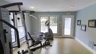 This single-floor fitness space is designed to support a well-rounded exercise routine in a clean, functional environment. The room is finished with cool grey walls and a textured ceiling, lending it a calm, focused atmosphere suited for physical activity. Large windows along the exterior wall flood the space with natural light and offer views of the outdoor area beyond, which includes a patio and surrounding greenery. A glass-paneled door provides direct access to the outdoors, enhancing ventilation and convenience. The flooring is a durable, light-toned surface appropriate for gym use. Equipment on hand includes a multi-function strength training rack, a weight bench, and an elliptical cross-trainer, offering options for both resistance and cardiovascular workouts. Recessed lighting supplements natural light, ensuring the space remains well-lit at any time of day. An emergency exit sign is prominently displayed, reflecting attention to safety standards. The layout is open and uncluttered, allowing for comfortable movement between equipment. Overall, this fitness room delivers a practical and inviting setting for daily exercise, combining essential amenities with a bright, airy feel.