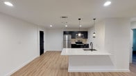 Located in Dallas's vibrant 75206 zip code, this single-floor residence showcases a clean, contemporary design with consistent finishes throughout. The open-concept living and kitchen area is anchored by a striking white quartz waterfall island with an undermount sink and matte black faucet. The kitchen is outfitted with dark navy cabinetry featuring brushed gold hardware, a decorative tile backsplash, and a full suite of stainless steel appliances including a refrigerator, range, and over-the-range microwave. Stylish pendant lights hang above the island, complementing the recessed lighting overhead. Light-toned hardwood-style flooring flows throughout most of the space, providing a warm contrast to the crisp white walls. Bold matte black interior doors add a modern edge and create a cohesive design statement visible throughout the hallway and common areas. The bedrooms are bright and well-proportioned, each featuring natural light from black-framed windows with outdoor views of greenery. The bathroom features large-format white wall tile, a soaking tub, penny tile flooring, and matte black fixtures, delivering a polished, modern aesthetic. Recessed lighting and clean lines unify the design of this well-appointed Dallas residence.
