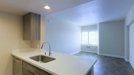 This single-floor space offers a thoughtfully designed open-concept layout that seamlessly connects the kitchen and living area. The kitchen is appointed with sleek white quartz countertops that extend into a peninsula, providing both prep space and a casual dining surface. A stainless steel undermount sink with a modern pull-down faucet serves as a functional centerpiece, complemented by warm wood-toned cabinetry that adds depth and contrast to the otherwise light and airy palette. Recessed lighting above the kitchen illuminates the workspace effectively. The adjacent living area is finished with dark flooring that grounds the space and creates a striking contrast against the neutral walls. A large window floods the room with natural light, enhancing the open and inviting atmosphere. The textured ceiling adds subtle visual interest throughout. The overall design is clean and contemporary, with a neutral color scheme that provides a versatile backdrop for a variety of interior styles. The flowing connection between the kitchen and living spaces makes this layout practical and well-suited for everyday living and entertaining alike.