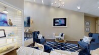 This single-floor space showcases a thoughtfully designed living area that blends classic elegance with modern comfort. The room immediately draws the eye upward with its vaulted ceilings, which lend a sense of openness and grandeur to the space. A decorative chandelier serves as a refined focal point overhead, complementing the warm, neutral tones of the walls.

The living area is anchored by a bold navy and cream color scheme, reflected in the plush seating arrangement that includes a tufted navy wingback chair, a cream settee, and a generously sized sofa. A geometric navy and white area rug ties the seating together, adding visual interest to the rich hardwood flooring beneath. A round metal coffee table with a tray display sits at the center, providing both functionality and style.

A wall-mounted flat-screen television is framed prominently on the main wall, flanked by recessed lighting that allows for customizable ambiance. An abstract navy canvas and a wall sconce add artistic touches, while a console table with candle accents contributes warmth and character. Track lighting along the ceiling further enhances the room's illumination. The overall atmosphere is polished, inviting, and well-appointed, making it an ideal space for both relaxing and entertaining.