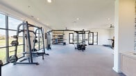 This generously sized fitness center offers a comprehensive workout environment thoughtfully designed to accommodate a wide range of exercise routines. The single-floor layout provides an open, unobstructed floor plan that promotes easy movement between functional zones. The flooring features durable rubber gym material with designated padded areas, offering both safety and comfort during training.

The strength training section is well-appointed with multiple cable machines, a functional trainer, free-weight racks, barbells, kettlebells, medicine balls, and adjustable benches — providing everything needed for a full resistance training regimen. A large wall mirror in one area enhances the sense of openness and allows for form monitoring during workouts.

The cardio zone features treadmills, elliptical trainers, stair climbers, and a rowing machine, positioned to take full advantage of the floor-to-ceiling grid-pane windows that frame stunning views of a resort-style outdoor pool and surrounding grounds. Abundant natural light pours throughout the space, creating an energizing atmosphere.

Overhead, large ceiling fans ensure consistent air circulation, while recessed lighting provides bright, even illumination. Black-framed glass doors add a sleek, modern aesthetic and connect different areas of the facility seamlessly. The clean white walls and modern design elements contribute to a professional yet welcoming fitness environment.