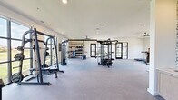 This generously sized fitness center offers a comprehensive workout environment thoughtfully designed to accommodate a wide range of exercise routines. The single-floor layout provides an open, unobstructed floor plan that promotes easy movement between functional zones. The flooring features durable rubber gym material with designated padded areas, offering both safety and comfort during training.

The strength training section is well-appointed with multiple cable machines, a functional trainer, free-weight racks, barbells, kettlebells, medicine balls, and adjustable benches — providing everything needed for a full resistance training regimen. A large wall mirror in one area enhances the sense of openness and allows for form monitoring during workouts.

The cardio zone features treadmills, elliptical trainers, stair climbers, and a rowing machine, positioned to take full advantage of the floor-to-ceiling grid-pane windows that frame stunning views of a resort-style outdoor pool and surrounding grounds. Abundant natural light pours throughout the space, creating an energizing atmosphere.

Overhead, large ceiling fans ensure consistent air circulation, while recessed lighting provides bright, even illumination. Black-framed glass doors add a sleek, modern aesthetic and connect different areas of the facility seamlessly. The clean white walls and modern design elements contribute to a professional yet welcoming fitness environment.