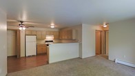 This single-floor space offers a practical and functional open-concept layout that connects the kitchen and living area in a cohesive, easy-to-navigate floor plan. The kitchen is outfitted with warm honey-toned wood cabinetry that provides ample upper and lower storage, complemented by a white refrigerator and a range. A built-in peninsula serves as a natural divider between the kitchen and the living area, offering additional counter space and the potential for casual seating. The kitchen features durable hardwood-style flooring, while the adjacent living area transitions to plush wall-to-wall carpeting, creating a comfortable and welcoming atmosphere. Ceiling-mounted light fixtures provide consistent ambient lighting throughout the space. A doorway visible from the living area suggests additional rooms beyond, indicating further functional living space. The neutral color palette of whites, beiges, and warm wood tones throughout lends the space a clean, understated aesthetic that is easy to personalize. Overall, the layout prioritizes practicality and open flow, making it well-suited for everyday living.
