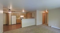This single-floor space offers a practical and functional open-concept layout that connects the kitchen and living area in a cohesive, easy-to-navigate floor plan. The kitchen is outfitted with warm honey-toned wood cabinetry that provides ample upper and lower storage, complemented by a white refrigerator and a range. A built-in peninsula serves as a natural divider between the kitchen and the living area, offering additional counter space and the potential for casual seating. The kitchen features durable hardwood-style flooring, while the adjacent living area transitions to plush wall-to-wall carpeting, creating a comfortable and welcoming atmosphere. Ceiling-mounted light fixtures provide consistent ambient lighting throughout the space. A doorway visible from the living area suggests additional rooms beyond, indicating further functional living space. The neutral color palette of whites, beiges, and warm wood tones throughout lends the space a clean, understated aesthetic that is easy to personalize. Overall, the layout prioritizes practicality and open flow, making it well-suited for everyday living.