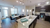 This single-floor residence showcases a thoughtfully designed open-concept layout that seamlessly connects the kitchen, dining, and living areas into one cohesive living space. The kitchen serves as the centerpiece, featuring a spacious granite-topped island with a double stainless steel sink, ideal for both meal preparation and casual entertaining. Dark espresso cabinetry provides a striking contrast against the light countertops, while sleek hardware and finishes add a polished, contemporary touch. Pendant lighting hangs elegantly above the island, complementing a track lighting system that ensures the space is well-lit throughout. The adjoining dining area is bathed in natural light from large windows with bright white blinds, creating a warm and inviting atmosphere. A ceiling fan overhead adds comfort and functionality. The hardwood-style flooring flows continuously throughout the open area, contributing to a unified and modern aesthetic. Neutral wall tones create a versatile backdrop that works harmoniously with the bold cabinetry and accents. The overall ambiance is clean, bright, and contemporary, making this space both functional and visually appealing for everyday living and entertaining.