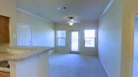This single-story space features an open living area that serves as the central hub of the layout. The room is finished with plush wall-to-wall carpeting and neutral-toned walls, creating a clean and versatile backdrop. Crown molding lines the ceiling perimeter, adding a refined architectural detail, while a ceiling fan with a light fixture provides both airflow and ambient lighting. Natural light enters generously through two windows fitted with horizontal blinds, flanking a glass-paneled exterior door that leads outside. The door and windows together create a bright, welcoming atmosphere even when closed. A ceiling-mounted HVAC vent ensures efficient climate control throughout the space. Adjacent to the living area, a partial view of a granite-topped counter suggests a kitchen or breakfast bar nearby, hinting at a convenient open-concept connection between the living and kitchen areas. The overall ambiance is bright, functional, and move-in ready, with classic details like crown molding and stone-look countertops elevating the space above standard finishes. The single-floor layout offers ease of accessibility and a cohesive flow throughout.