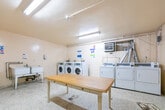 Located at 1470 Williamsport Rd in Mississauga, Ontario, this shared laundry facility provides a practical and functional space designed to serve the needs of its users efficiently. The room is finished with durable terrazzo flooring and smooth, light-coloured walls that contribute to a clean, utilitarian atmosphere. Overhead fluorescent lighting ensures the space is well-lit throughout.

The laundry area is equipped with multiple front-loading washing machines and top-loading dryers arranged along the walls, offering ample capacity for regular use. A utility sink is also present, providing added convenience for hand-washing or pre-treating garments. A central wooden folding table serves as a practical surface for sorting and folding laundry.

Ventilation infrastructure is visible along the upper walls, supporting the dryer exhaust systems. Informational notices and usage guidelines are posted on the walls, helping users operate the equipment properly. The space is accessed through two separate doors, allowing for straightforward entry and exit. A wall-mounted unit provides additional climate control within the room.

Overall, this laundry facility is a well-equipped, no-frills communal space that prioritizes functionality and accessibility, making it a convenient amenity for residents at this Mississauga address.