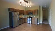 This single-floor residence offers a functional and inviting layout designed for comfortable everyday living. The kitchen serves as a central highlight, showcasing a full suite of stainless steel appliances, including a refrigerator, range with oven, built-in microwave, and dishwasher. Rich, warm-toned wood cabinetry lines the walls, providing generous storage space, while granite-look countertops add a polished finishing touch. A stainless steel sink is positioned beneath upper cabinets, maximizing workspace efficiency. The flooring throughout features durable hardwood-style planks in a warm, natural tone that seamlessly ties the space together. Overhead, a combination of track lighting and a ceiling fan ensures both ample illumination and air circulation. The walls are finished in a soft, muted green hue that complements the cabinetry and flooring, creating a cohesive and welcoming atmosphere. The open sightlines from the kitchen into the adjacent living area enhance the sense of space, making the layout feel airy and connected. Overall, this residence balances practical design with a warm, approachable aesthetic suited for a variety of lifestyles.