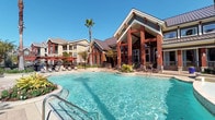 This single-story amenity facility offers a thoughtfully designed environment that blends resort-style outdoor living with polished interior spaces. At the heart of the outdoor area is a large, sparkling swimming pool with crystal-clear turquoise water, framed by ample poolside seating, pergola-shaded lounging areas, and lush landscaping including a signature palm tree. The surrounding architecture features bold red-wood columns, a steeply pitched roof, and warm earth-toned exteriors that lend the space a welcoming, upscale character set against an open blue sky.

Inside, the interiors are finished with natural stone tile flooring in a soft, neutral palette that carries through the corridors and entry areas. The entry hallway is accented with a large framed landscape artwork, a wall-mounted shelf, and subtle coat hooks, creating a functional yet aesthetically considered arrival experience. A large decorative mirror with a dark, richly finished frame adds depth and elegance to the interior corridor. Wall-mounted sculptural décor in textured bronze tones contributes warmth and visual interest throughout.

The facility also includes tanning bed stations, housed behind wire-front enclosures for ventilation, offering a practical wellness amenity. The overall ambiance is one of comfort and sophistication, designed to provide residents with a resort-like experience within a community setting.