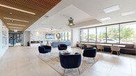 Located at 770 Brookfield Rd in Ottawa, Ontario, this expansive single-floor amenity space offers a thoughtfully designed, open-concept layout that blends sophisticated style with functional versatility. The space is defined by a striking slatted wood ceiling detail that adds warmth and texture, complementing the large-format porcelain tile flooring throughout. Floor-to-ceiling windows flood the interior with abundant natural light and provide views of the surrounding greenery.

The main lounge area serves as the social heart of the space, featuring a dramatic marble-clad fireplace wall with an integrated flat-screen display, plush sectional seating, and a statement sputnik chandelier. Adjacent to it, a refined seating area with deep navy velvet chairs and a dark tiled accent wall with open shelving creates an inviting atmosphere. A bold "Hello Ottawa" mural adds a vibrant, locally inspired focal point.

The collaborative zone offers generous natural wood communal tables beneath contemporary pendant lighting, ideal for group work or casual dining. A cozy library nook with built-in bookshelves, a neon sign, and a living green wall adds a creative retreat. The entry corridor features suspended circular swing chairs and a stainless steel elevator core clad in dark stone tile, completing this well-appointed, design-forward space.