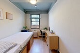 Located at 419 W 34th St in the heart of Midtown Manhattan, this efficiently designed private room offers a well-organized layout that maximizes every inch of its compact footprint. The room features light-toned wood flooring throughout, which contributes to a clean and bright aesthetic. A distinctive muted teal ceiling accent adds a subtle design element that sets the space apart from a purely utilitarian look.

The bedroom is thoughtfully furnished with a platform bed frame equipped with built-in storage drawers underneath, providing practical space-saving solutions. Adjacent to the bed, a warm wood-toned built-in desk and credenza unit offers ample workspace and storage, making it well-suited for studying or working. A wall-mounted mirror above the credenza adds depth to the room.

Natural light enters through a window fitted with a window-unit air conditioner and a radiator below, ensuring year-round climate comfort. The window offers a glimpse of surrounding greenery. A compact refrigerator and small plant add functional and decorative touches.

The en-suite area, positioned near the entry door, includes a wall-mounted sink with a mirror cabinet and a shower with a curtain. A closet provides additional storage. The overall design is practical and tidy, well-suited for single occupancy in one of New York City's most central and accessible neighborhoods.
