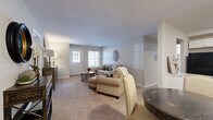 This single-floor residence offers a thoughtfully organized layout comprising three bedrooms, three bathrooms, a living room, kitchen, hallway, and two closets. The living room serves as the heart of the home, featuring neutral-toned carpet, soft natural light from a front-facing entry door with a windowed panel and an adjacent window, and understated wall decor that creates a calm, inviting atmosphere. The hallway connecting the rooms is accented with warm wood-tone flooring and provides an elegant transition space, as seen with decorative console furnishings and artwork along its walls. The kitchen is efficiently designed with white shaker-style cabinetry, granite-look countertops, wood-look flooring, and black appliances including a refrigerator, range, microwave, and dishwasher. The primary bedroom offers a generous footprint with plush carpet, soft neutral walls, and recessed lighting. The additional bedrooms, also carpeted, provide flexible use — one notably accommodating a home office setup with a window for natural light. The bathrooms are clean and functional, featuring white vanities with ample counter space, wood-look tile flooring, and tub-shower combinations. Two closets provide convenient storage throughout the residence. Overall, the space maintains a cohesive, neutral palette that is both practical and easy to personalize.