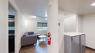 This thoughtfully designed single-floor unit makes excellent use of its compact footprint, offering a functional layout that includes a combined kitchen and living area, one bedroom, a hallway, and a full bathroom. The open-plan living and kitchen space features warm wood-look flooring, clean white walls, and a panel ceiling with track lighting. A gray sectional sofa and round coffee table anchor the living area, which receives natural light through a window fitted with roller shades. The kitchen is defined by a breakfast bar with two black stools, gray cabinetry, a stainless steel range, microwave, and refrigerator — all neatly arranged for efficient use. The bedroom is a standout feature of the unit, equipped with a striking floor-to-ceiling dark metal perforated wardrobe system that doubles as a room divider and storage solution, paired with a built-in platform bed with under-bed storage and a compact desk workspace. The bathroom offers a clean, all-white tub and shower surround with a minimalist aesthetic. Throughout the unit, the consistent use of neutral tones, wood flooring, and modern fixtures creates a cohesive, contemporary ambiance well-suited to efficient urban living.