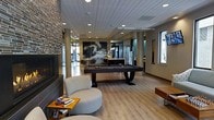 This single-floor amenity lounge is designed to offer a dynamic and social atmosphere through a thoughtfully curated mix of recreational and relaxation features. The open-plan layout is anchored by a sleek, dark-finished pool table positioned at the center of the space, making it an immediate focal point upon entry. Light hardwood flooring runs throughout, complementing the contemporary aesthetic and contributing to a bright, welcoming ambiance.

A floor-to-ceiling stone-clad fireplace lines one wall, adding a warm and textural contrast to the otherwise modern interior. Adjacent to it, a curved upholstered ottoman and small side table stacked with books create an inviting casual seating area. Recessed lighting in the white paneled ceiling provides consistent, even illumination across the space.

A striking large-scale wave mural serves as a dramatic backdrop, adding artistic character and depth to the room. Structural columns punctuate the open floor plan, while windows along the perimeter allow natural light to filter in. Toward the rear, partially enclosed areas with glass partitions suggest private or semi-private spaces, potentially for meetings or additional amenities, enhancing the overall versatility of the lounge.