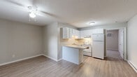 Located at 3150 Boardwalk Ln in Greenville, NC, this single-floor, one-bedroom, one-bathroom unit offers a practical and well-organized layout. The open-concept living, dining, and kitchen area creates a seamless flow, anchored by a peninsula-style kitchen with white cabinetry, light countertops, a double sink, and white appliances including an electric range and full-size refrigerator. Under-cabinet lighting and textured ceilings add character throughout the space. Light wood-look flooring runs through the living and kitchen areas, complementing the neutral gray walls and white trim. A ceiling fan with light fixture in the living area provides added comfort. The generously sized bedroom features plush carpeting, a ceiling fan, a closet with built-in rod and shelf, and a window with blinds that allows natural light to filter in. The bathroom is clean and functional, offering a tub-shower combination with tile surround, a single vanity, and wood-look flooring. Additional highlights include a dedicated laundry area, a utility room, and an exterior brick facade. The unit's efficient design makes thoughtful use of every square foot.