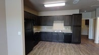 This single-floor space showcases a thoughtfully designed kitchen as its centerpiece. The kitchen features an L-shaped layout with rich, dark espresso-toned cabinetry that extends from floor to ceiling, providing generous storage throughout. The countertops display a light granite-look finish that creates a striking contrast against the dark cabinetry. A full suite of black appliances is included, featuring a refrigerator with freezer, a range with oven, and a dishwasher, offering a cohesive and practical setup. The double-basin sink is positioned along the back wall, complementing the overall functional layout. The flooring throughout consists of wood-look planks in a warm, medium-toned finish, adding texture and warmth to the space. Overhead, a flush-mounted fluorescent light fixture provides ample illumination across the kitchen workspace. The walls are finished in a neutral beige tone, contributing to a clean and versatile backdrop. The overall ambiance is modern and practical, with the dark cabinetry and black appliances lending a bold, contemporary feel to the interior.